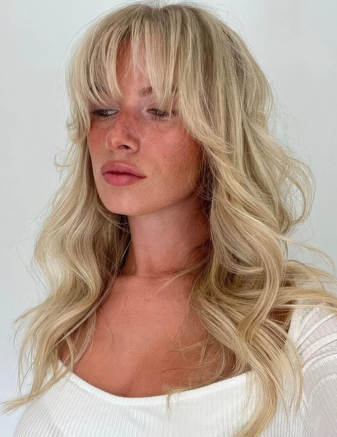 Wavy blonde hairstyle with a lot of dimension and Bardot bangs.