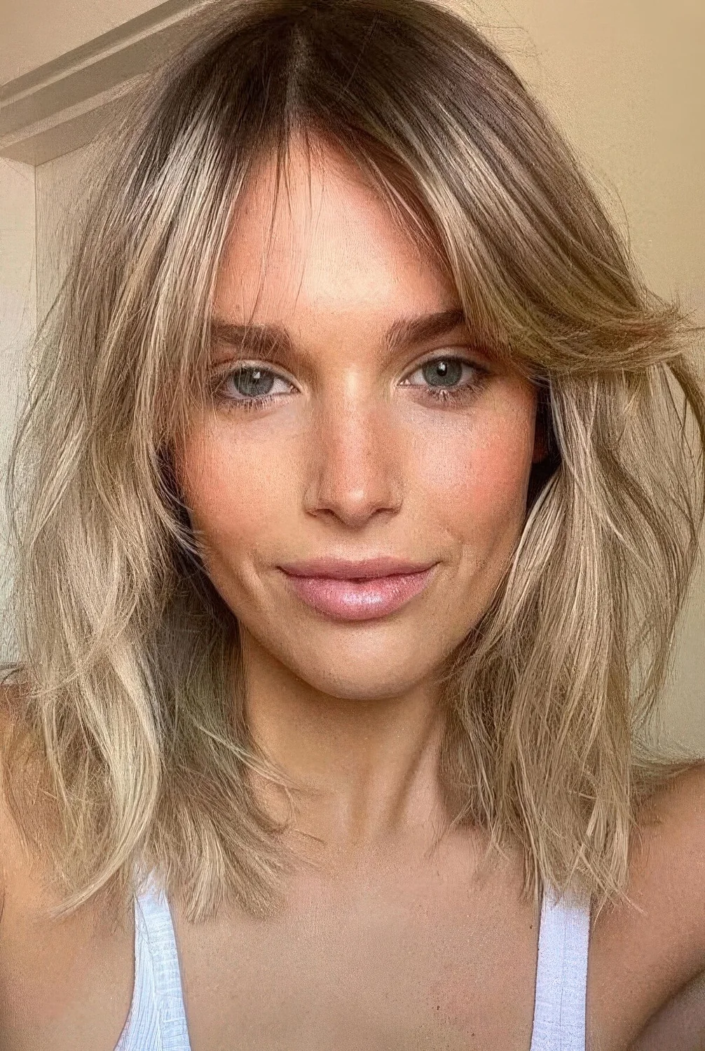 Long and messy blonde bob with a darker root and side bangs, with a lot of dimensional texture.