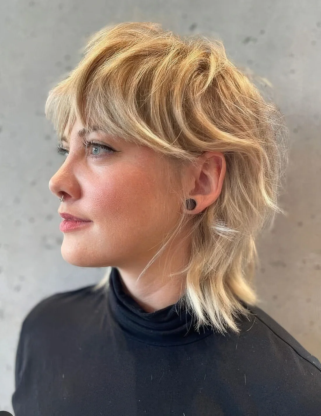 Textural mullet in blonde shades, with bangs, longer hair on top and shorter hair on the sides and back.