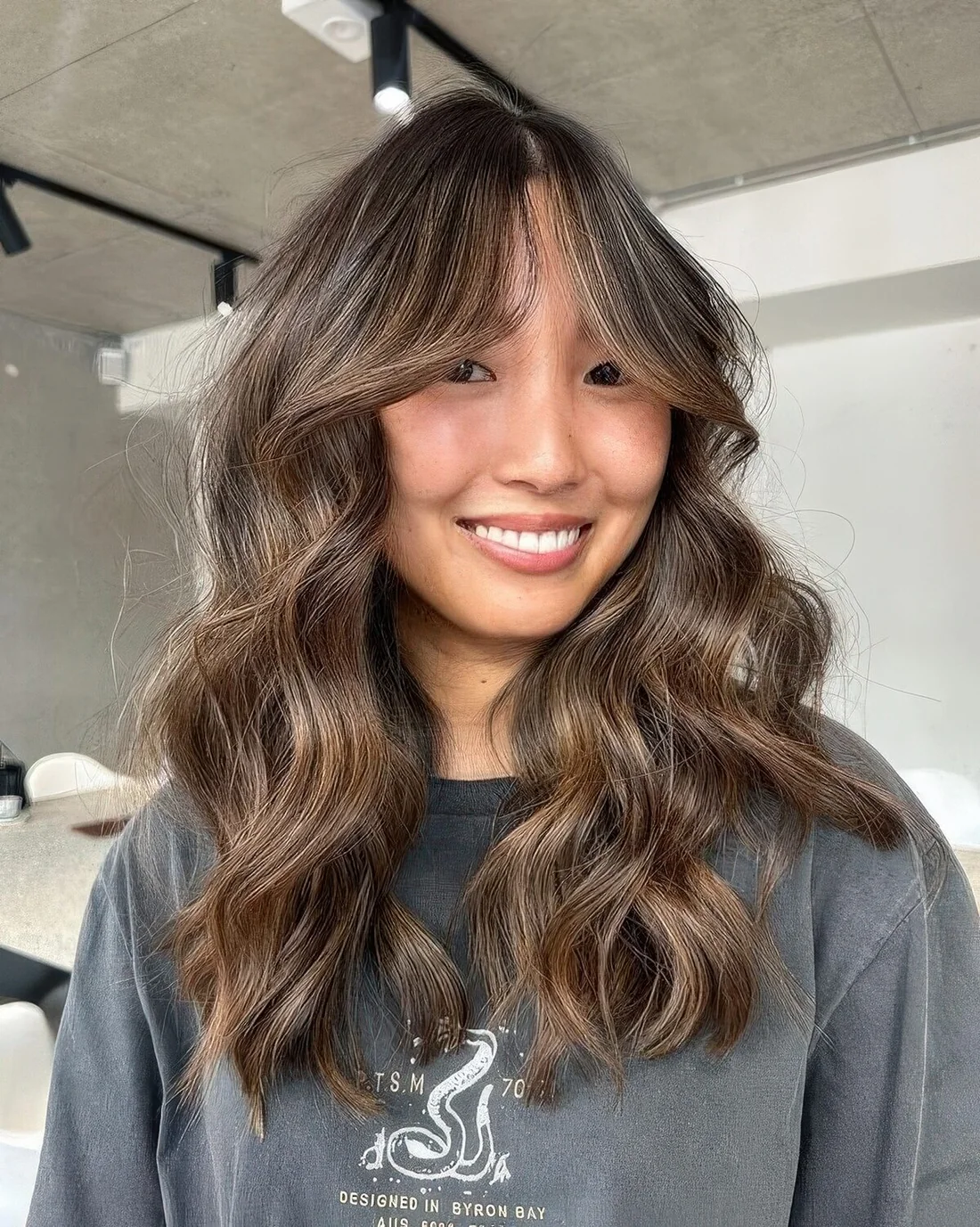 Long brunette butterfly haircut with caramel and bronde balayage with bottleneck bangs and waves.