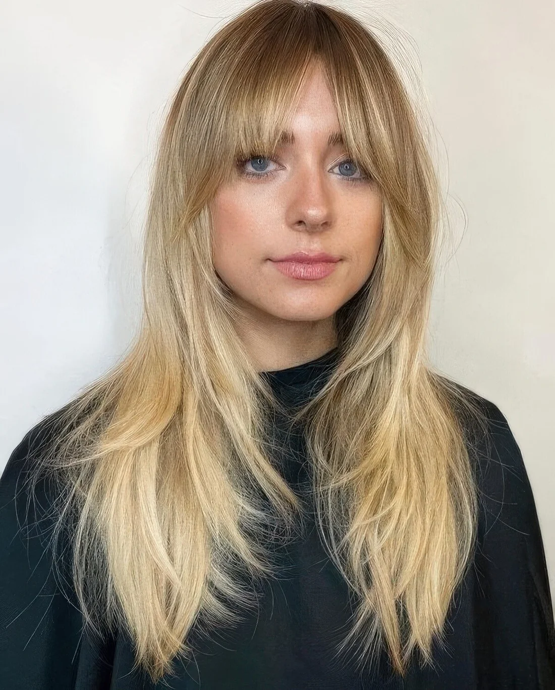 Long layered blonde haircut with bottleneck and curtain bangs.