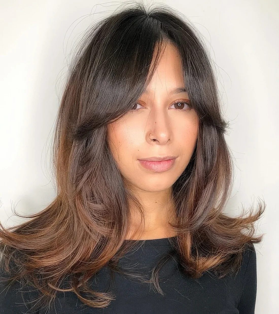 Dark brunette medium butterfly haircut with caramel balayage, bottleneck bangs and curled ends.