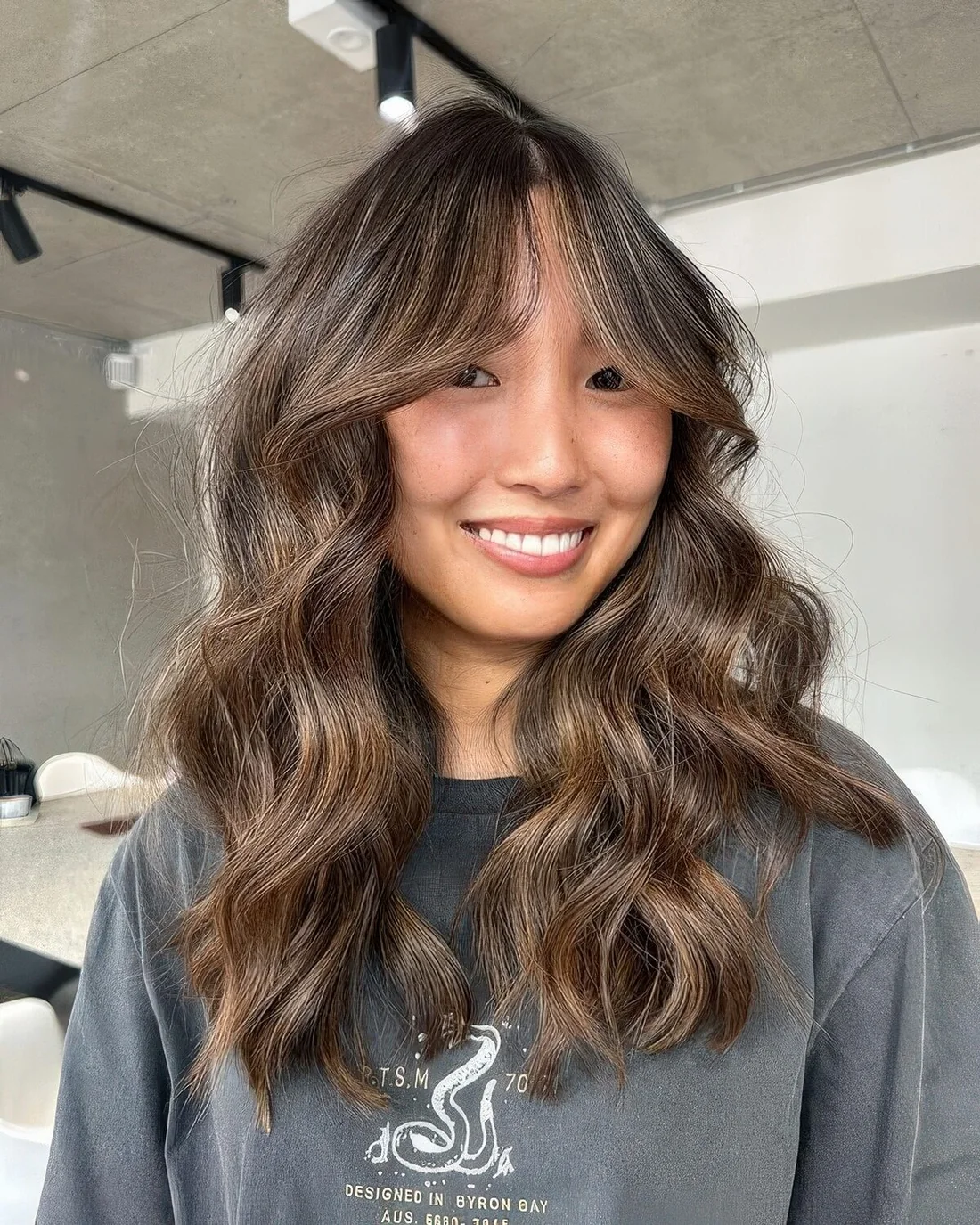 Long brunette butterfly haircut with caramel and bronde balayage with bottleneck bangs and waves.