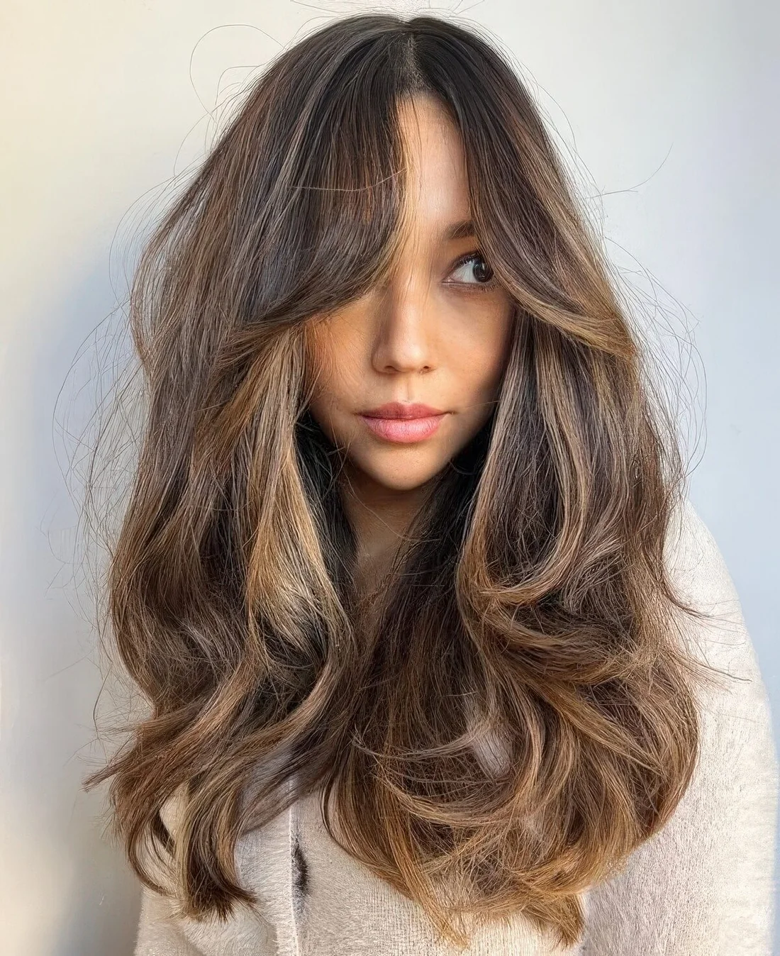 Long dark brunette hairstyle with caramel and golden blonde balayage, with bottleneck bangs and a lot of dimension and waves.