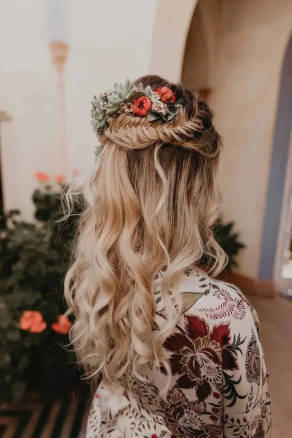 Boho wedding hairstyle with a fishtail braid halo, waves down and bold blooms and pale leaves tucked in.