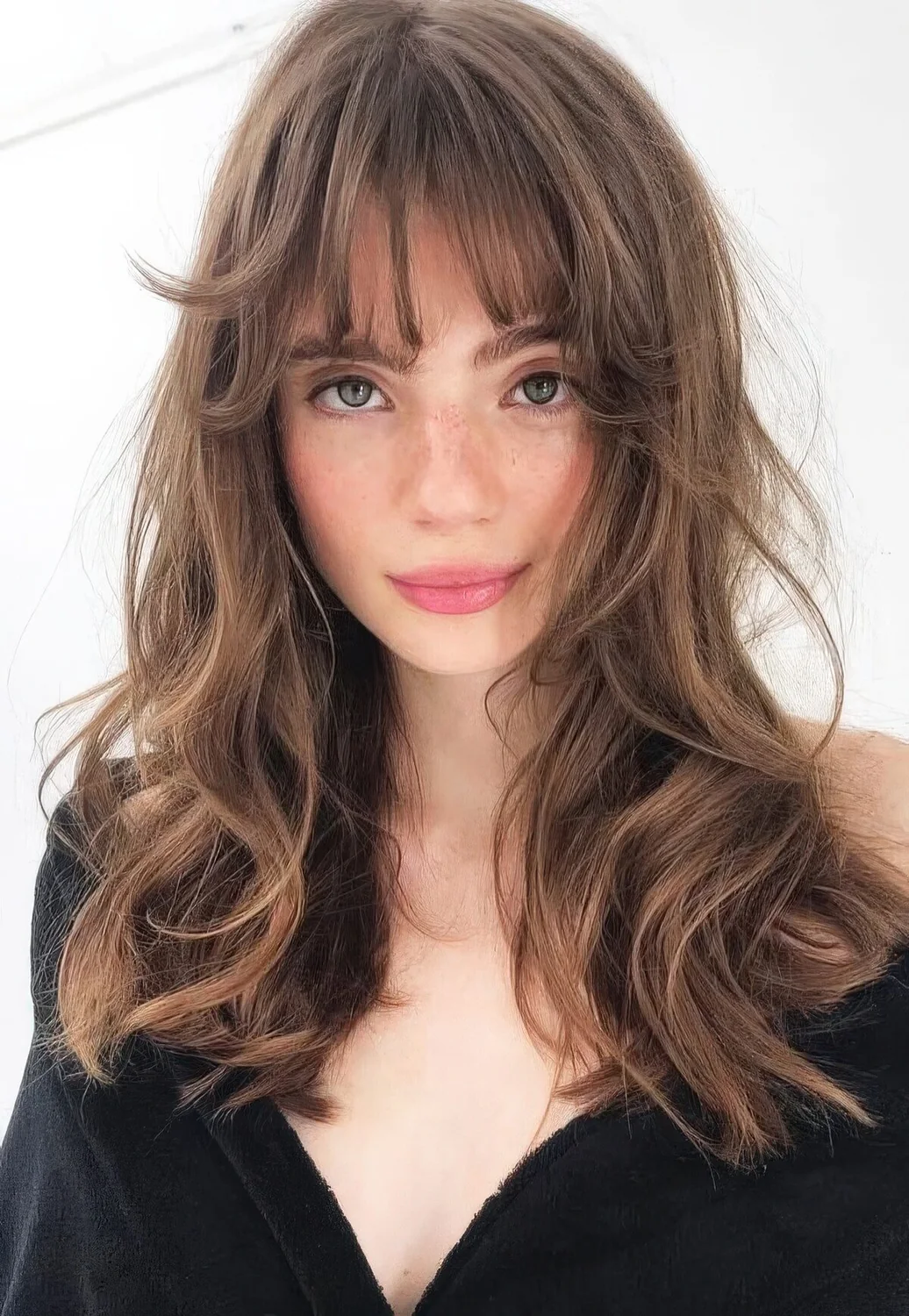 Long brunette butterfly haircut with messy waves and wispy bangs.