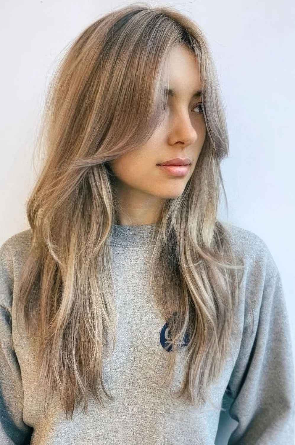 Long mushroom blonde hair with a slight balayage and long curtain bangs that perfectly frame the face.