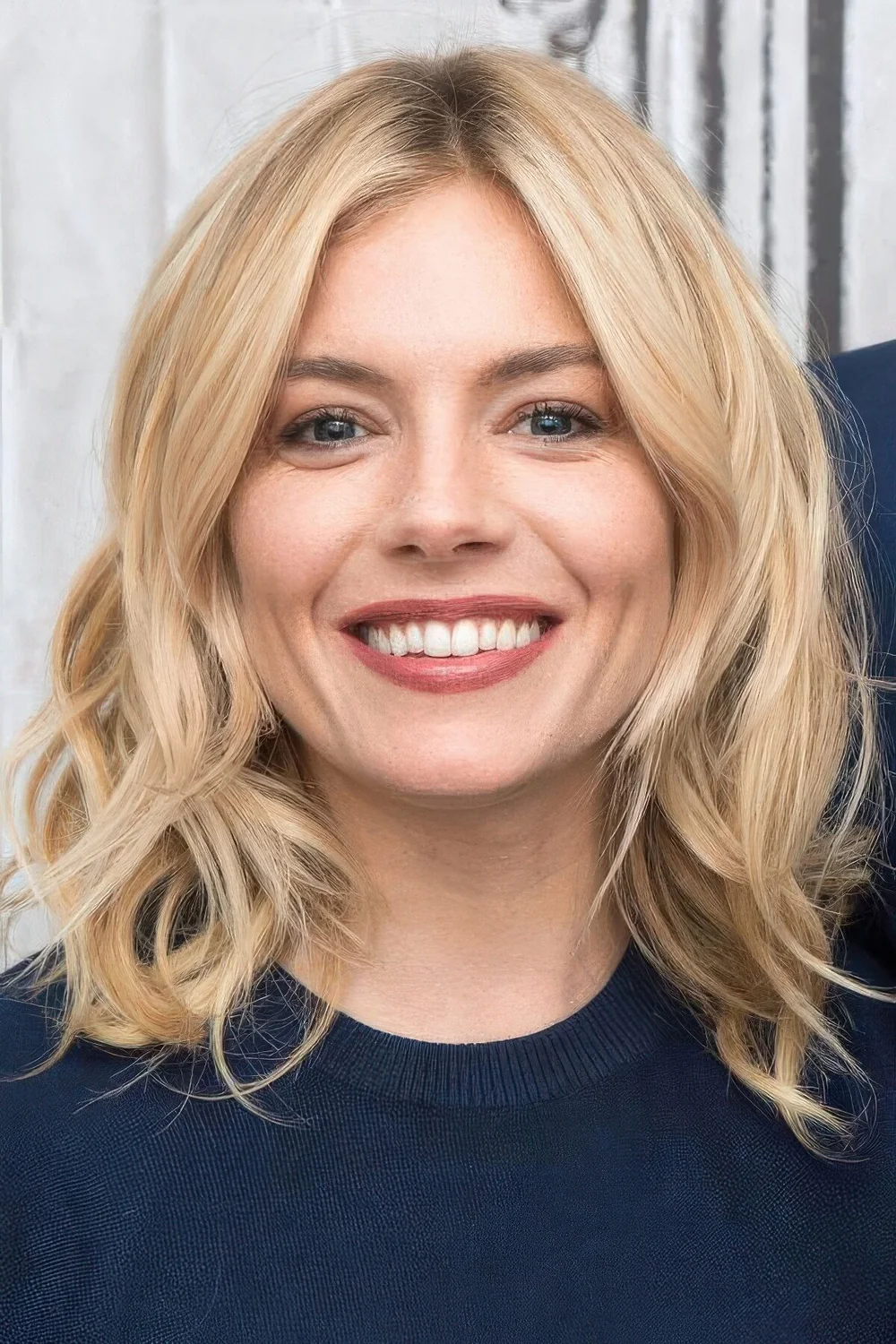 Sienna Miller rocking wavy blonde medium-length hair with chin bangs and waves.