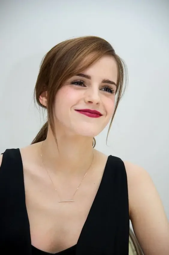 Emma Watson rocking light brunette hair in a low ponytail and side bangs.