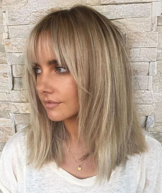 Medium-length layered haircut, blonde balayage, a darker root, layered bangs curtain and wispy ones.