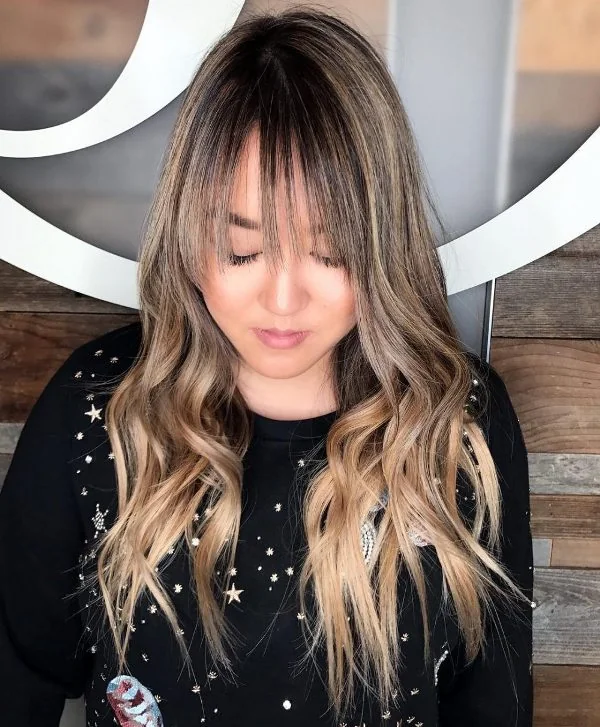 Long caramel blonde hair with darker lowlights and a dark root plus waves and wispy bangs for a touch of romance.