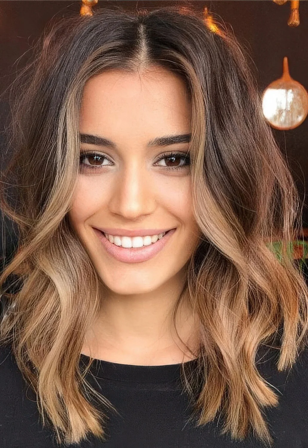 Volumetric dark brunette medium-length hair with caramel balayage and face-framing highlights plus waves.