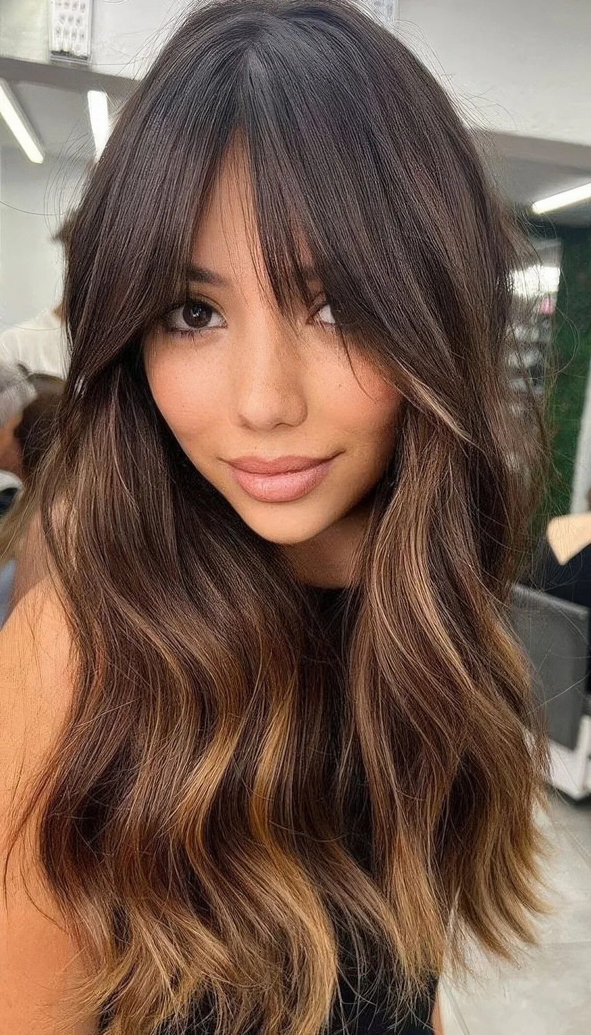 Long dark brunette hair with caramel highlights and bottleneck bangs.