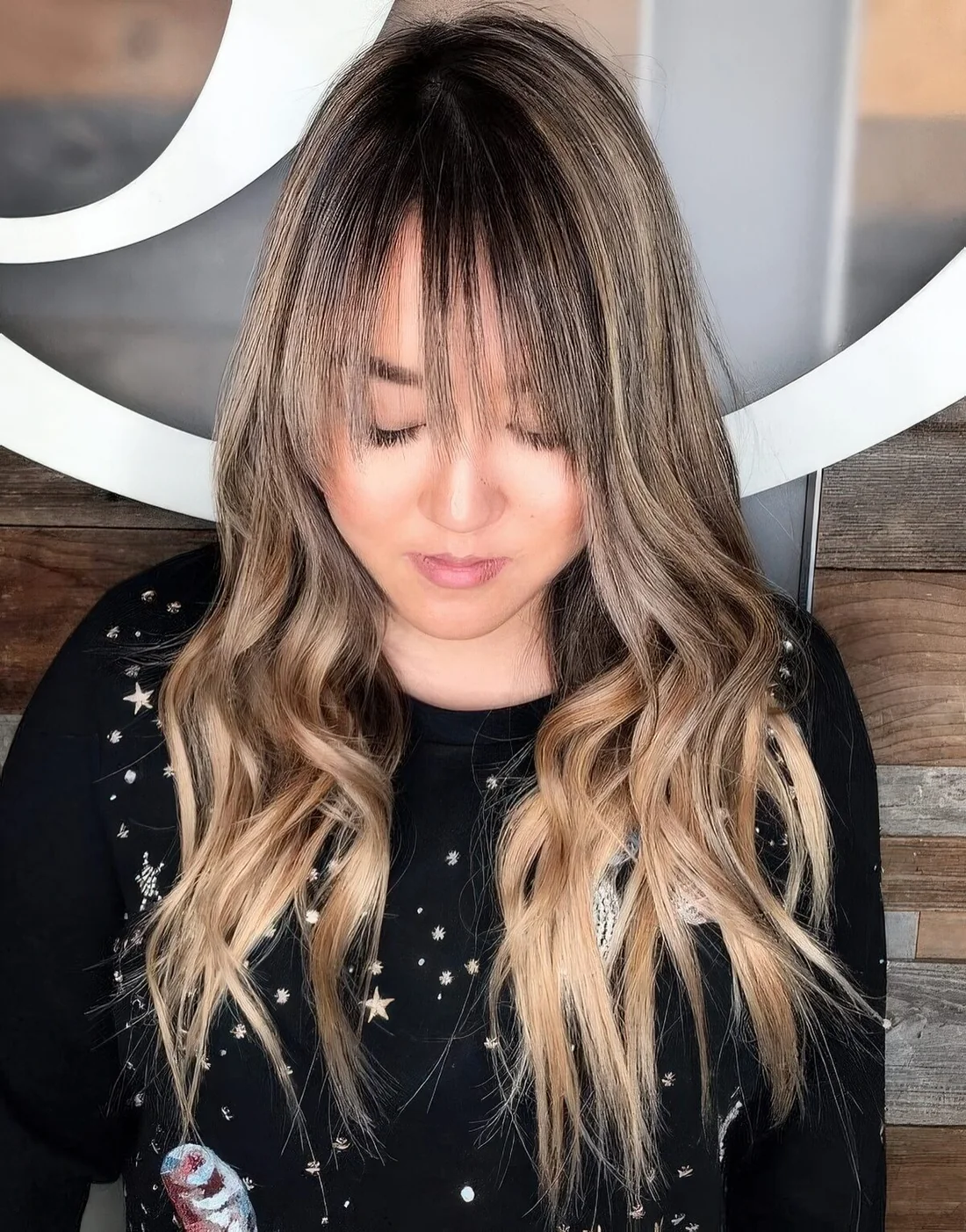 Long caramel blonde hair with darker lowlights and a dark root plus waves and wispy bangs for a touch of romance.