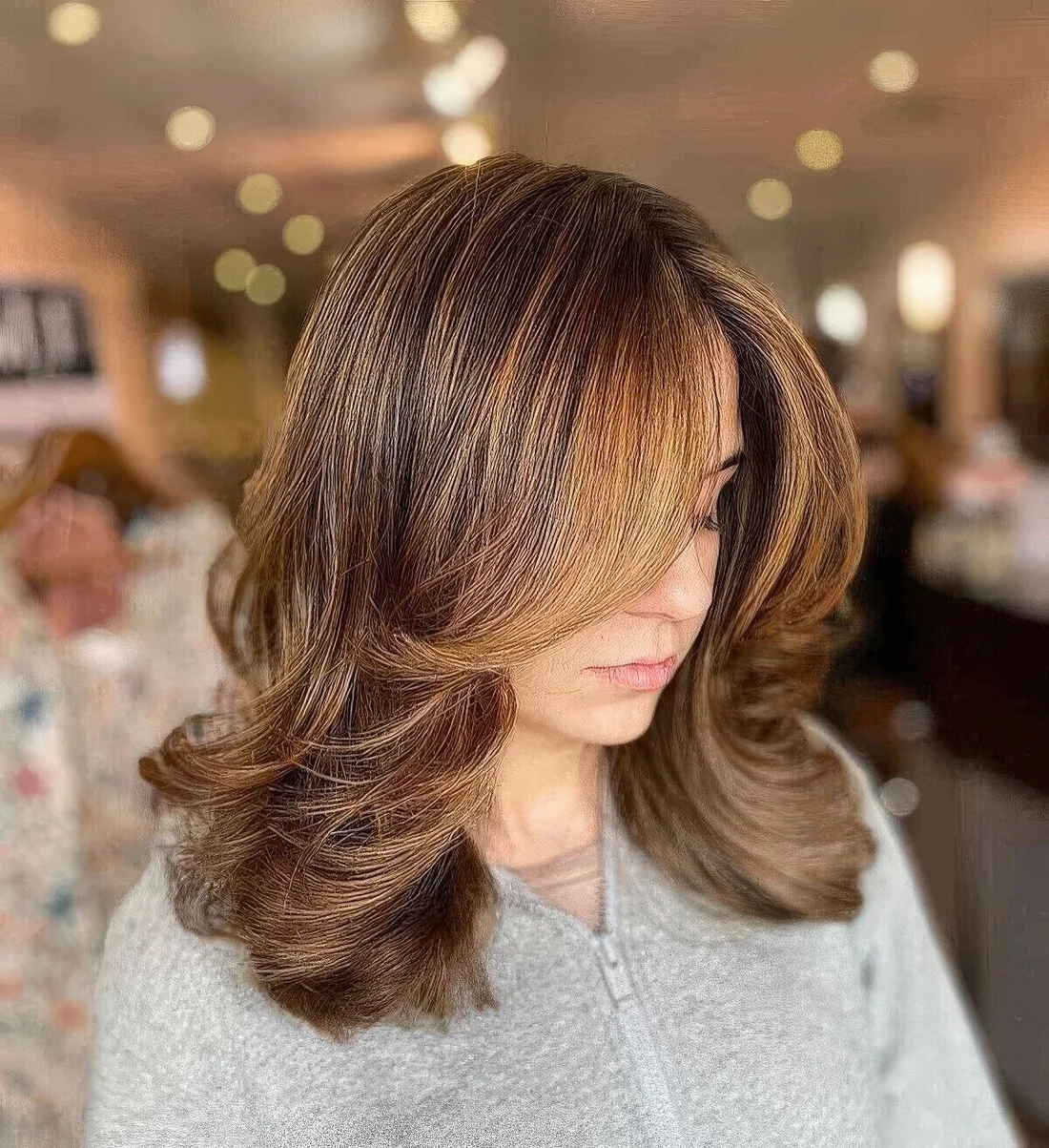 Chic brunette medium-length butterfly haircut with honey balayage and curtain bangs, a lot of volume and waved ends.