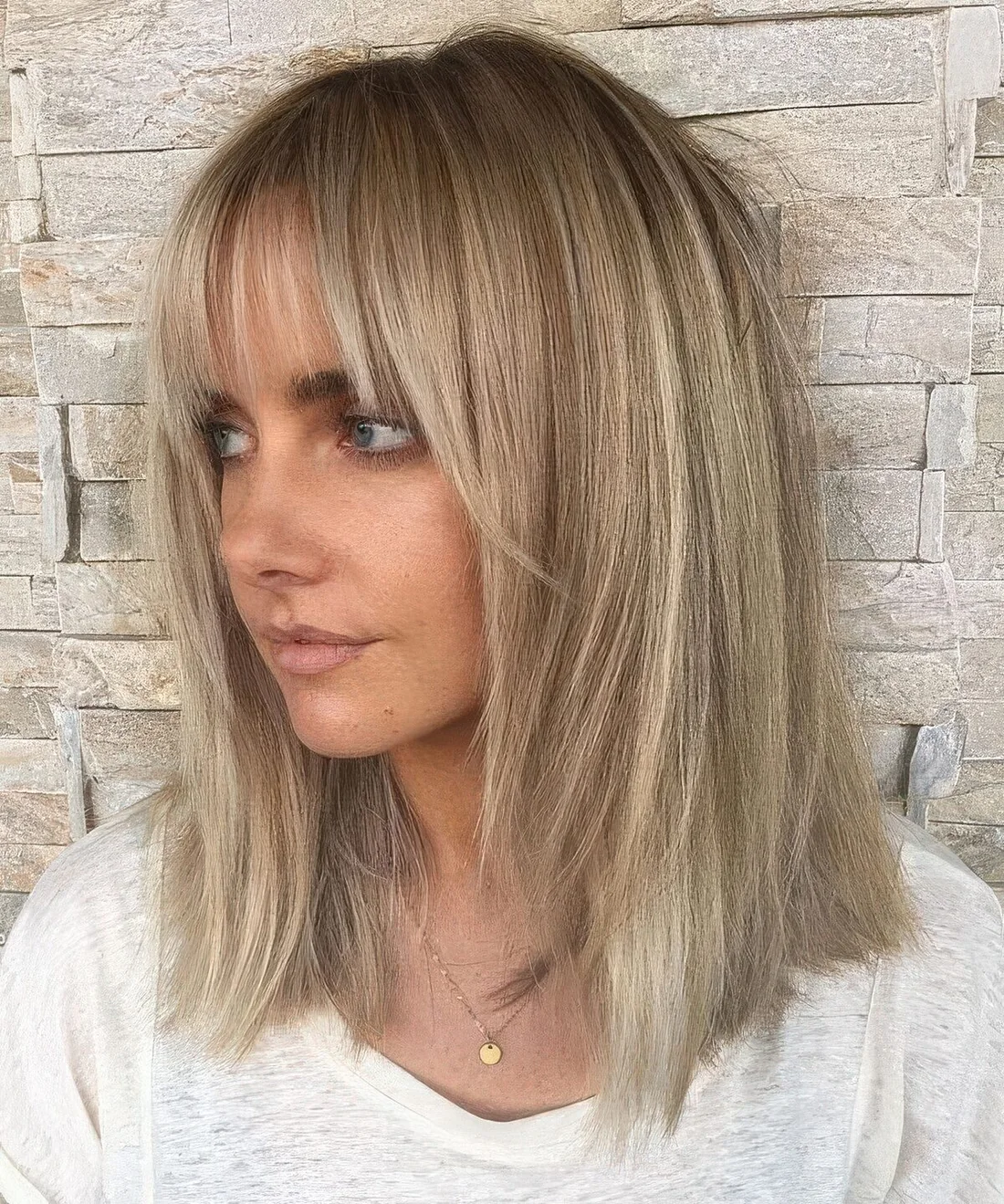 Medium-length layered haircut, blonde balayage, a darker root, layered bangs curtain and wispy ones.