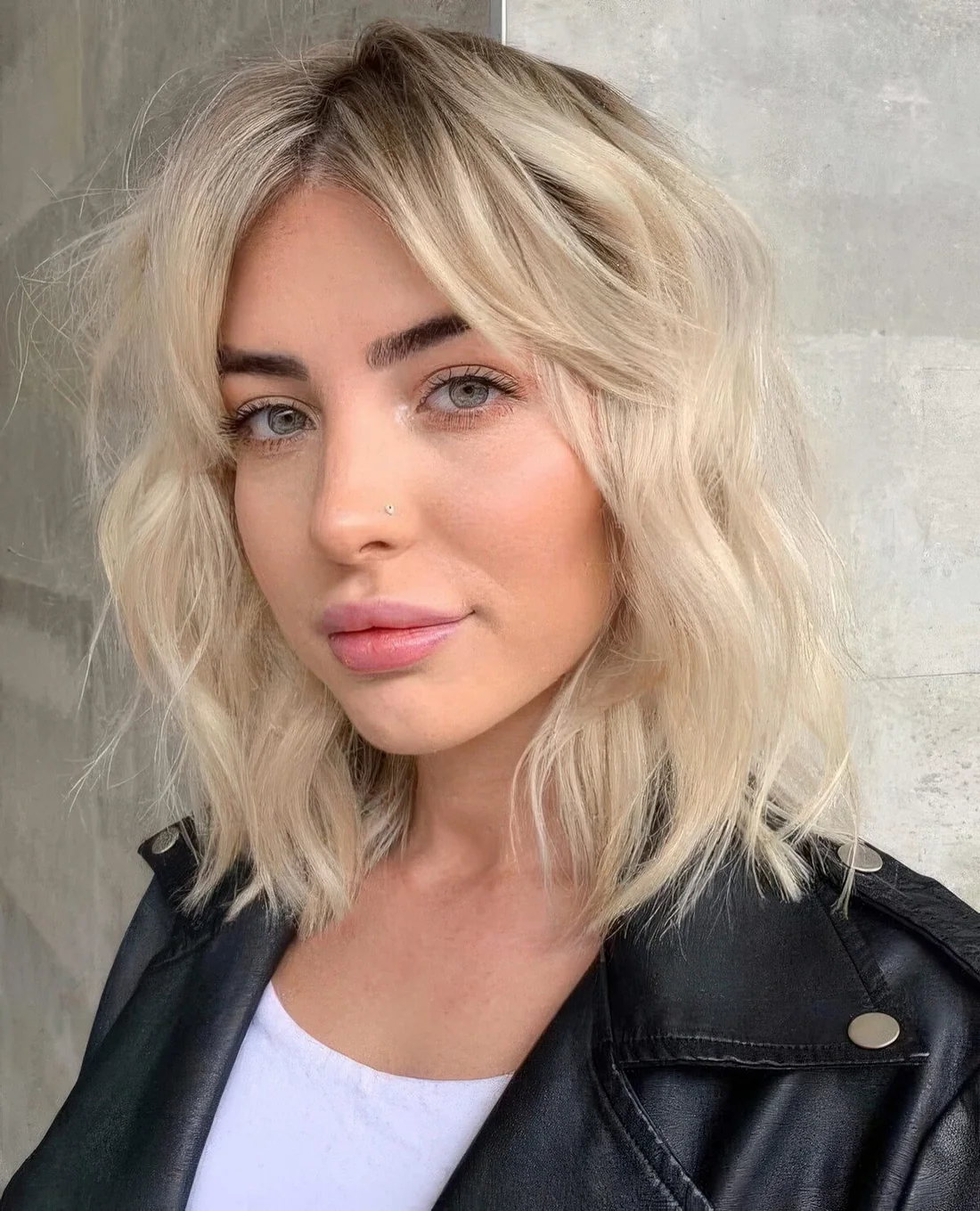 Bleached blonde shoulder-length hairstyle with messy and wavy volume with a dark root.