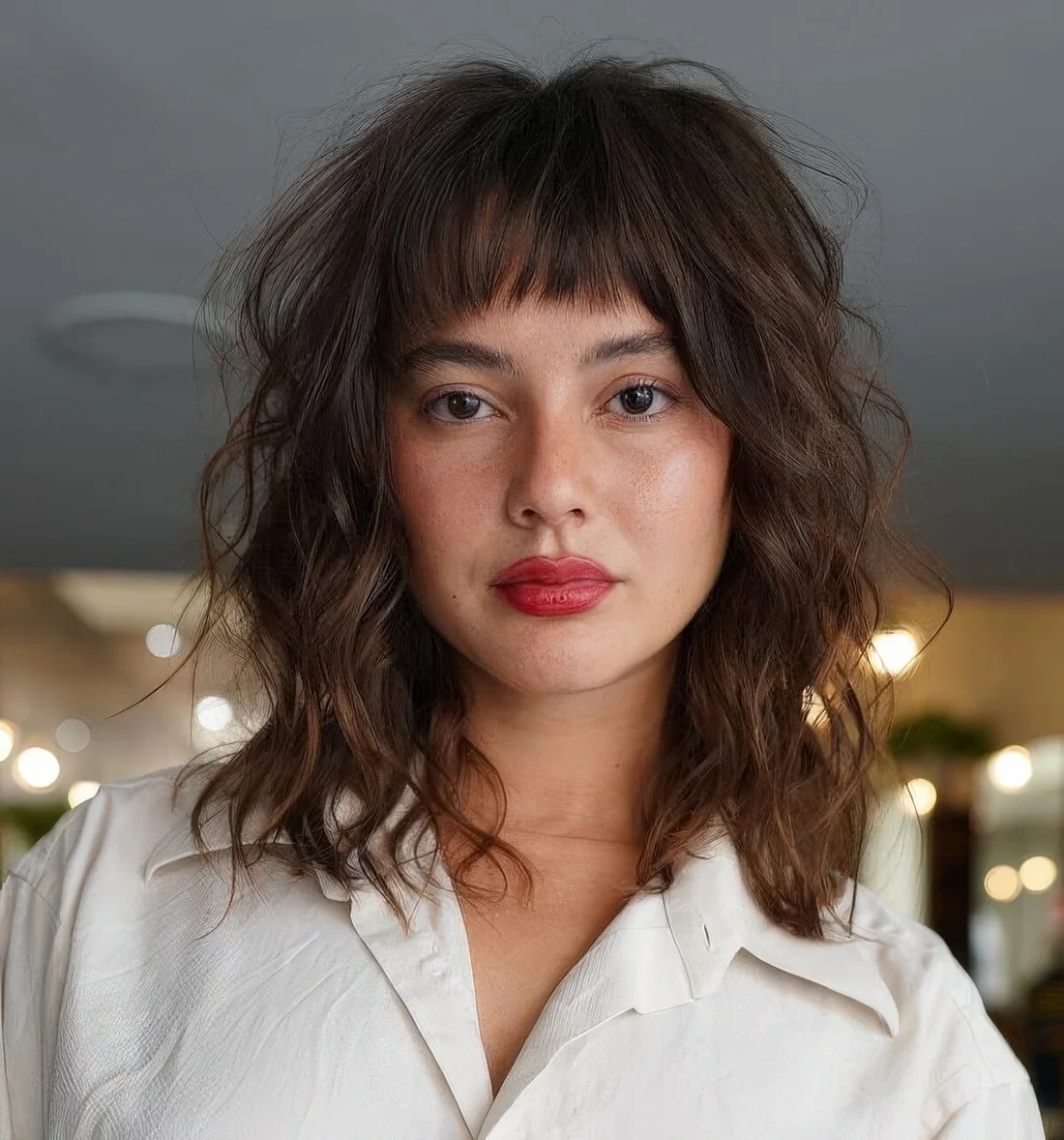 Dark brunette medium-length wavy hairstyle with highlights and blunt bangs.