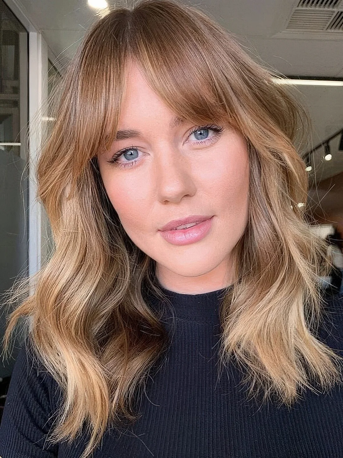 Long layered haircut in soft blonde shades, with short curtain bangs and messy waves to give a texture to the hair.