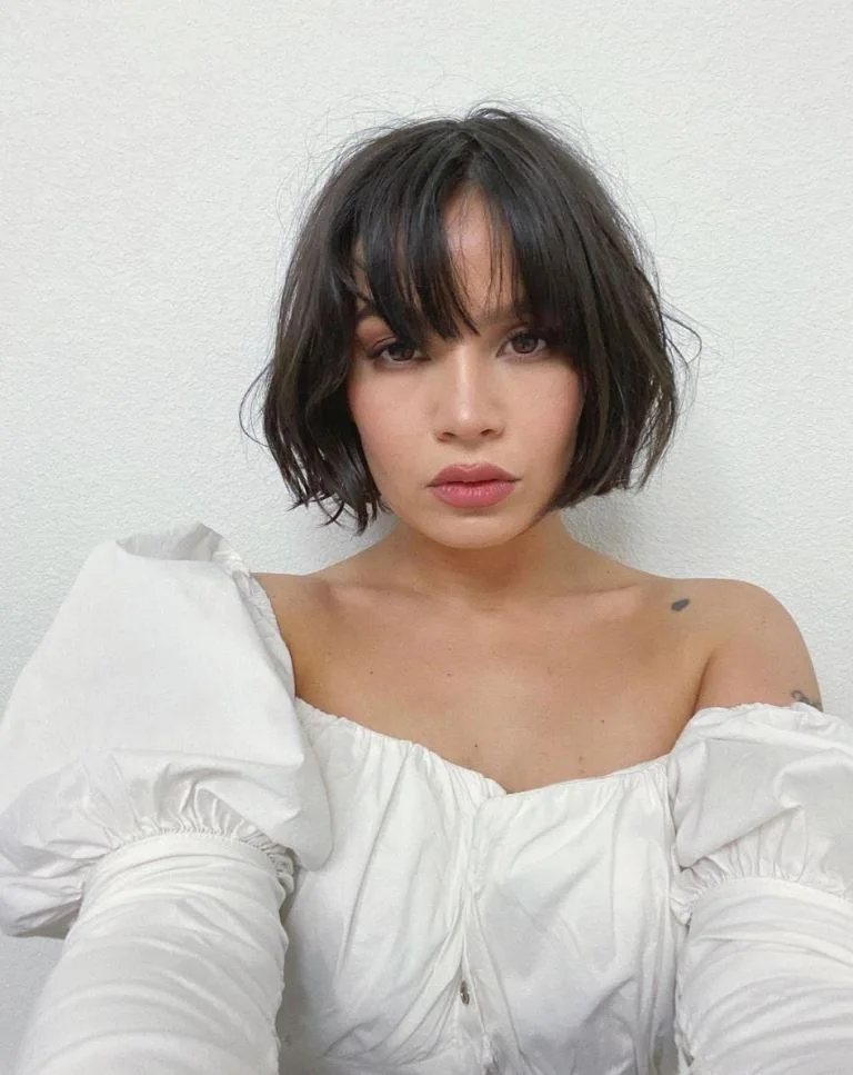 A black jaw length bob has loose waves, an off center part, and long bangs falling around the eyes