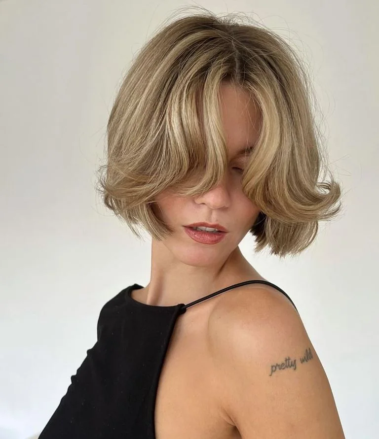 A short blonde bob features long curtain fringe, a center part, and softly curved ends