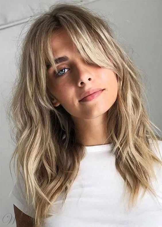 Blonde textural long hair with waves and layered bangs.