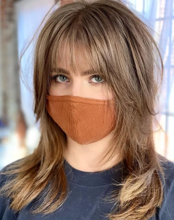 Long light brown shaggy hair with blonde balayage and lowlights, with layered bangs to make the look softer.