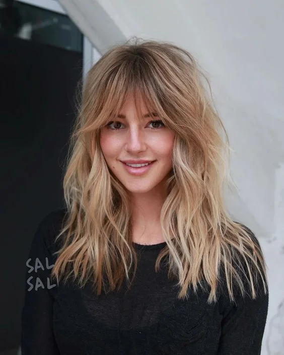 Long wavy blonde hair with plenty of texture and volume plus caramel balayage and layered bangs.