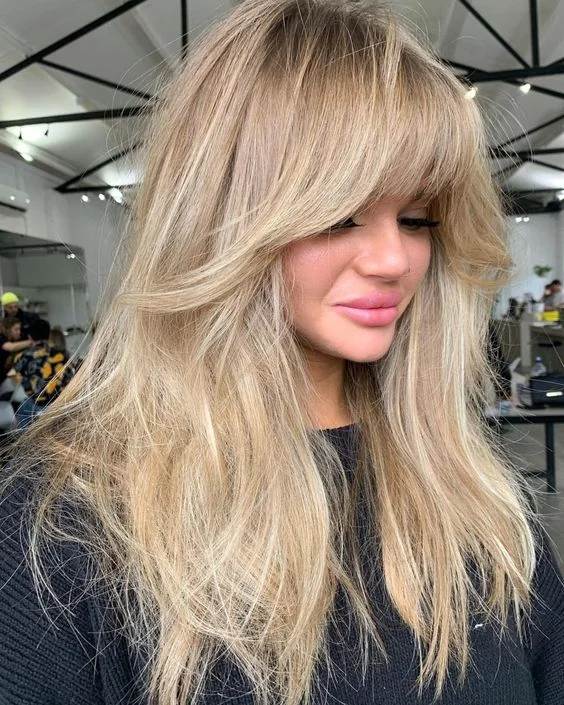 Naturally blonde long hair with balayage and much volume, with layered bangs and lowlights.