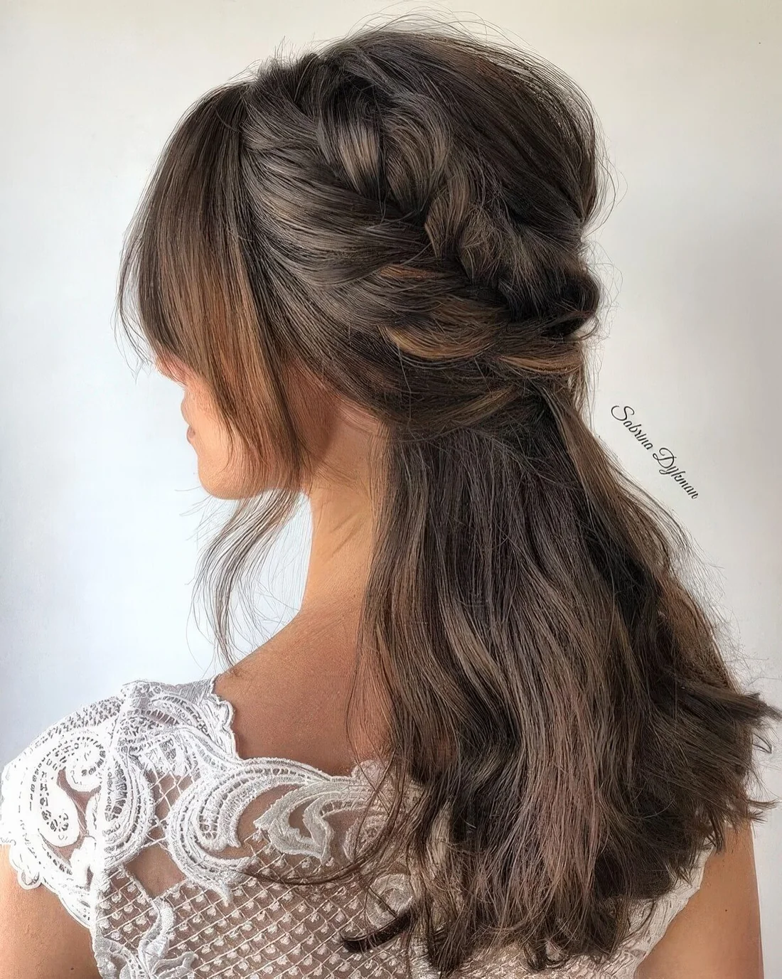 Half updo with a bump on top and a loose braided halo, face-framing bangs and textured hair down for a wedding.