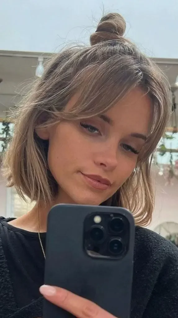 A blonde bob with face-framing fringe is gathered into a small top knot above the crown