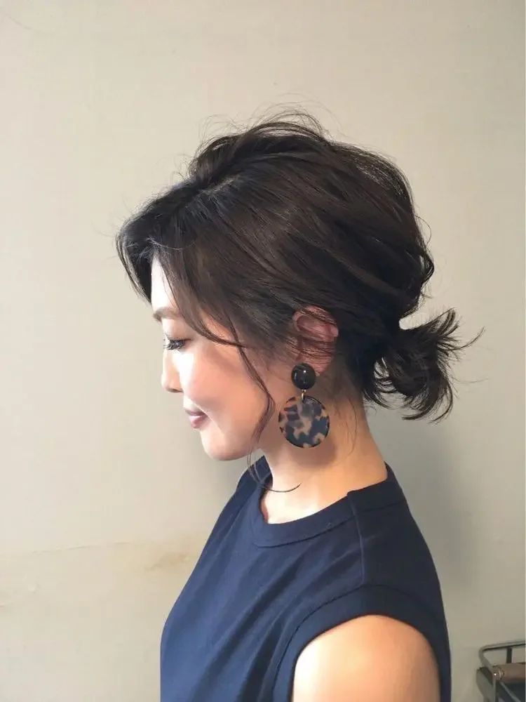 A dark wavy bob is gathered into a loose low ponytail with soft volume through the crown