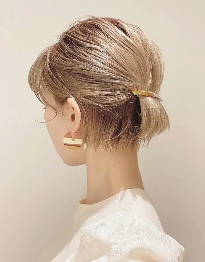 A short blonde bob is pulled into a tiny ponytail at mid-back with wispy pieces left loose